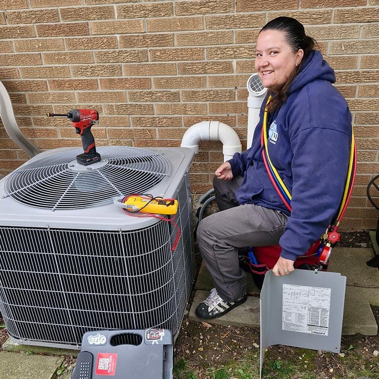 female D&M tech working on an exterior ac unit, sitting on a bucket, drill and multimeter tool on top of ac unit,