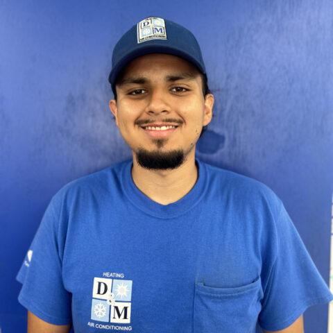 A man wearing a blue cap and blue T-shirt, both with DM Heating & Air Conditioning logos, stands smiling in front of a solid blue background.