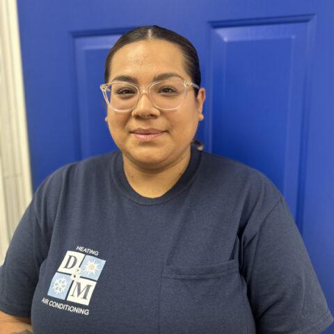 A person wearing glasses and a navy blue D&M Air Conditioning t-shirt stands in front of a bright blue door, smiling softly at the camera.