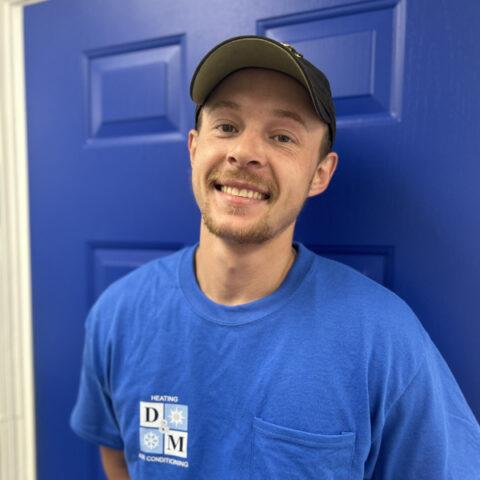 A smiling man wearing a black cap and a blue T-shirt with a DM Heating & Air Conditioning logo stands in front of a blue door.