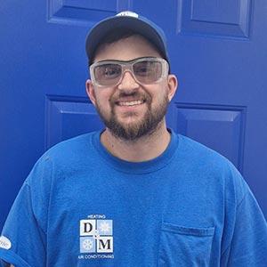 A man with a beard and glasses, wearing a blue cap and blue shirt with a DM Heating & Air Conditioning logo, stands smiling in front of a bright blue door.